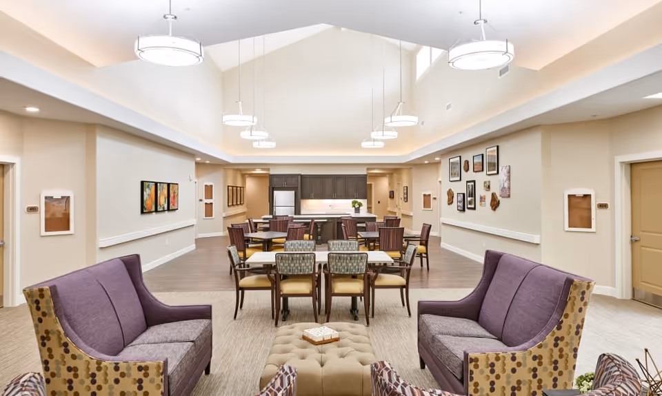 A spacious, well-lit common area in a senior living facility featuring two purple sofas with patterned sides facing each other, a tufted ottoman in the center, and several dining tables with chairs in the background. The room has high ceilings with multiple hanging light fixtures, beige walls adorned with framed artwork, and a kitchen area with dark cabinets and a refrigerator at the far end.