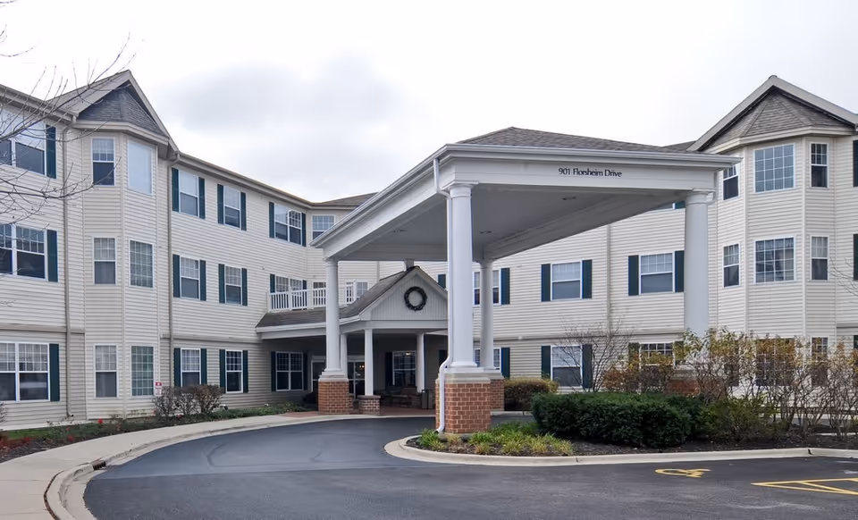 Exterior view of a three-story senior living facility building with beige siding and green shutters. The entrance features a covered driveway supported by white columns with brick bases. There are shrubs and small trees around the building, and the address 901 Florsheim Drive is visible on the entrance canopy.