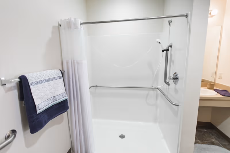 Accessible walk-in shower with grab bars, a handheld showerhead, a white curtain, and towels on a wall rack beside a vanity.