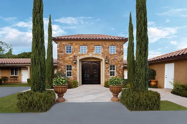 Front exterior view of a building with a stone facade, a tiled roof, and a large double door entrance flanked by two lantern-style lights. There are tall, narrow evergreen trees and large potted plants with flowers on either side of the entrance. The sky is partly cloudy.