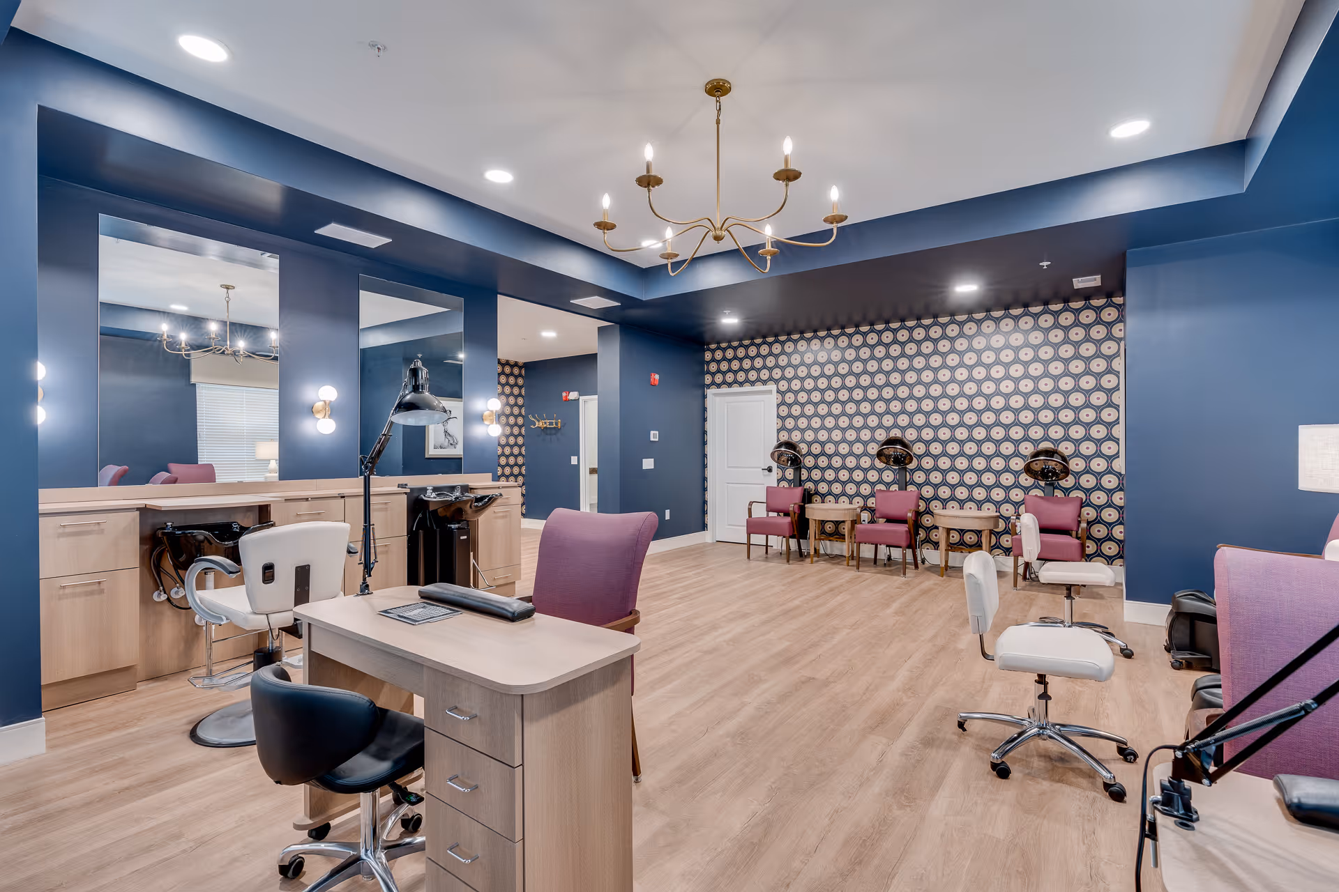 A spacious salon area in a senior living facility with blue walls and wood flooring. The room features multiple styling stations with mirrors, chairs, and hair washing sinks. There are several purple and white chairs, a chandelier hanging from the ceiling, and a patterned accent wall with hair drying chairs lined up against it.