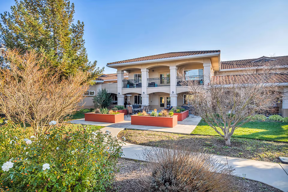 Outdoor view of The Meadows of Napa Valley facility showing a two-story building with a tiled roof, balconies, and a patio area with seating surrounded by landscaped garden beds and various trees and shrubs under a clear blue sky.