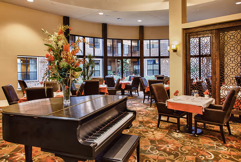 A dining room with multiple tables covered in white and orange tablecloths, surrounded by brown upholstered chairs. A black grand piano with a vase of colorful flowers on top is in the foreground. Large windows in the background show an outdoor courtyard area.