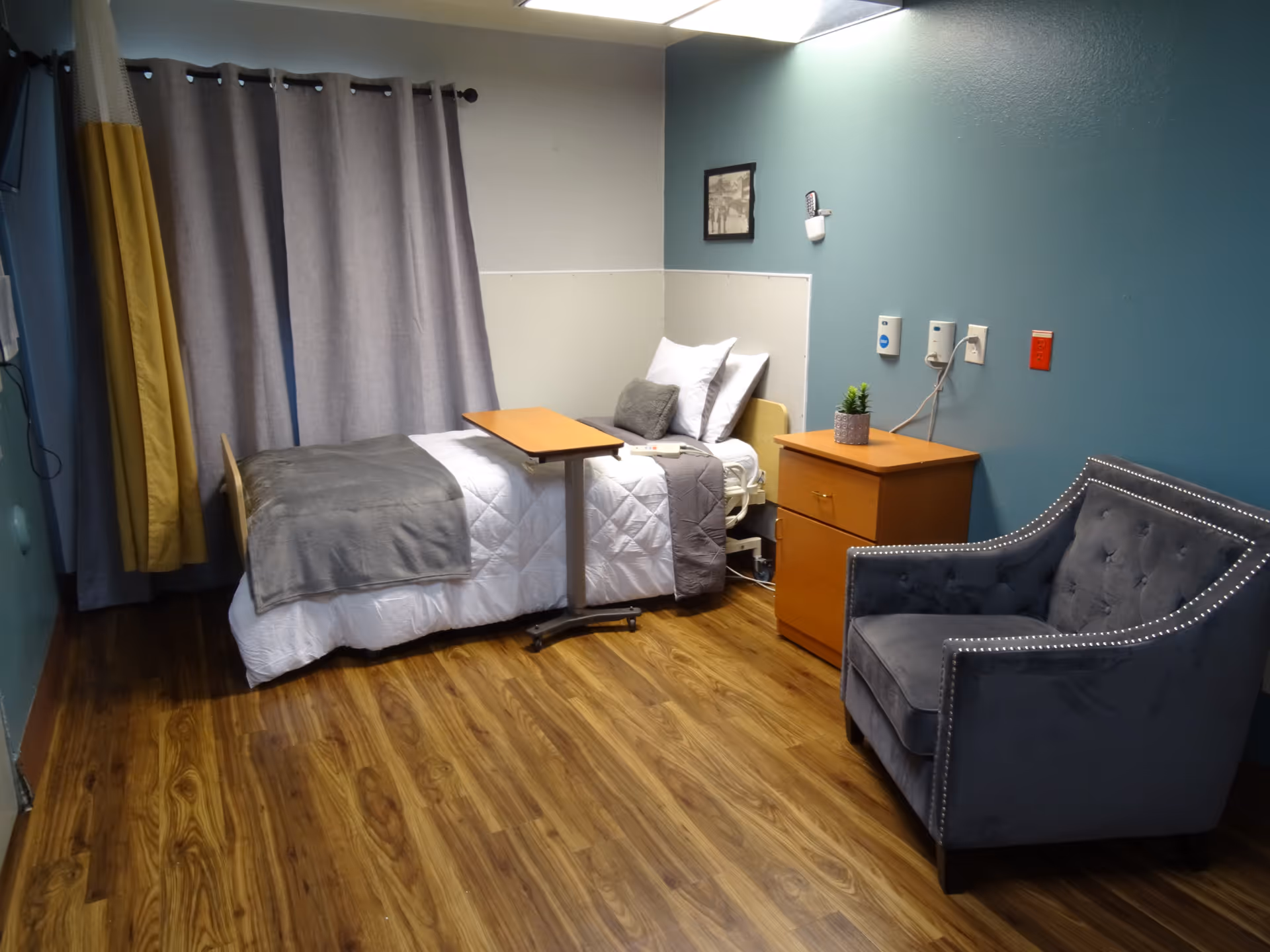 A cozy senior care room with a single bed made with white and gray bedding, a wooden overbed table, a wooden nightstand with a small plant, and a gray upholstered armchair. The walls are painted blue and white, and there are gray and yellow curtains covering the window. The floor is wood laminate.