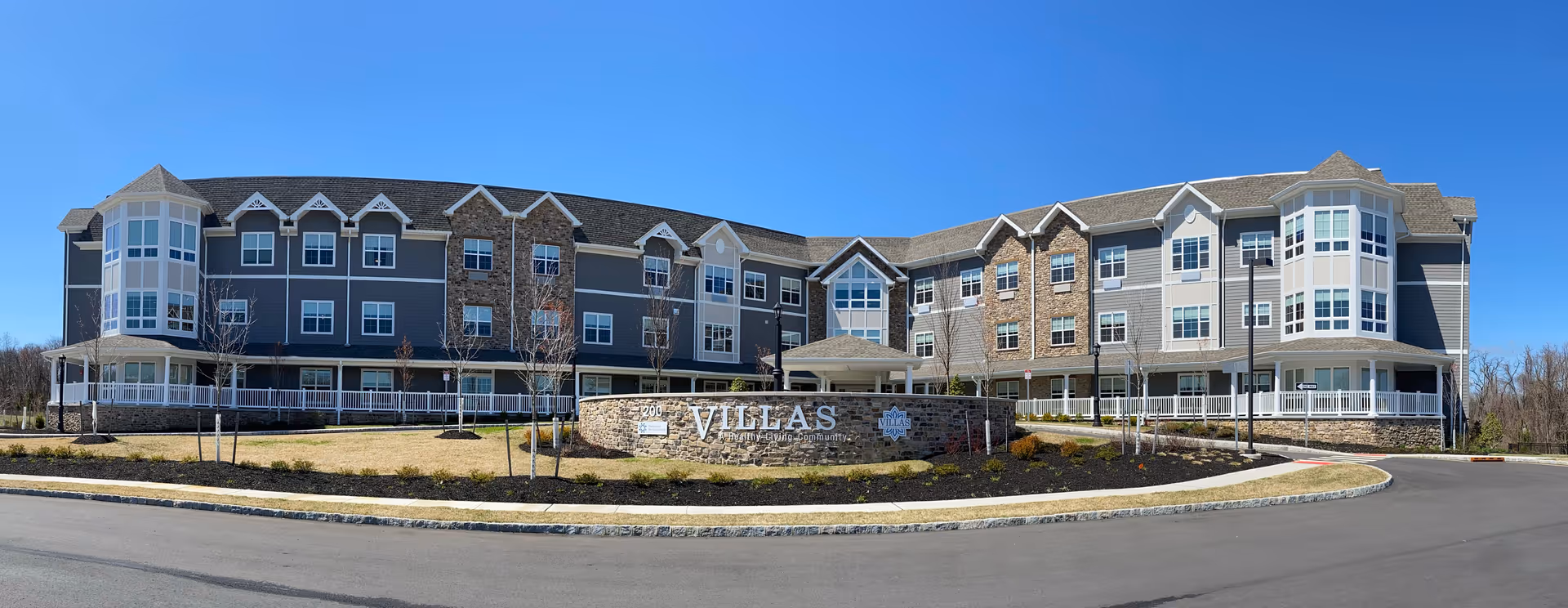 Wide exterior view of the Villas of Holmdel senior living facility building under a clear blue sky, featuring multiple windows, two prominent rounded corner sections with large windows, and a stone sign in front displaying the facility name.