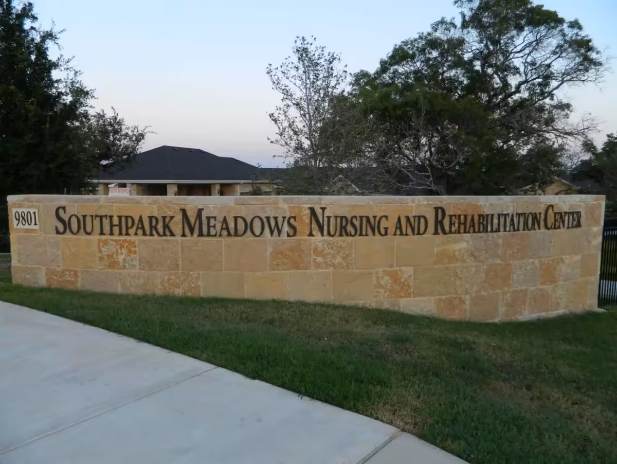 Stone sign with the text 'Southpark Meadows Nursing and Rehabilitation Center' on a grassy area next to a sidewalk, with trees and a building roof in the background.