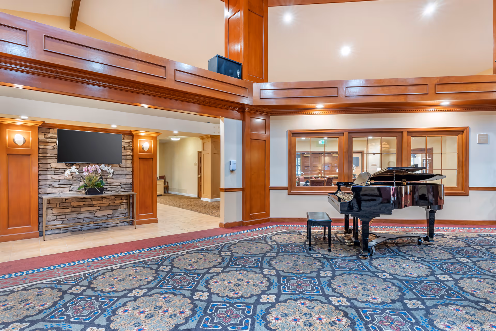 Interior view of a senior living facility common area featuring a black grand piano with a matching bench on a patterned carpet. The room has wooden paneling, a stone wall with a mounted flat-screen TV, and recessed lighting. There are windows looking into another room and a hallway leading to other areas.