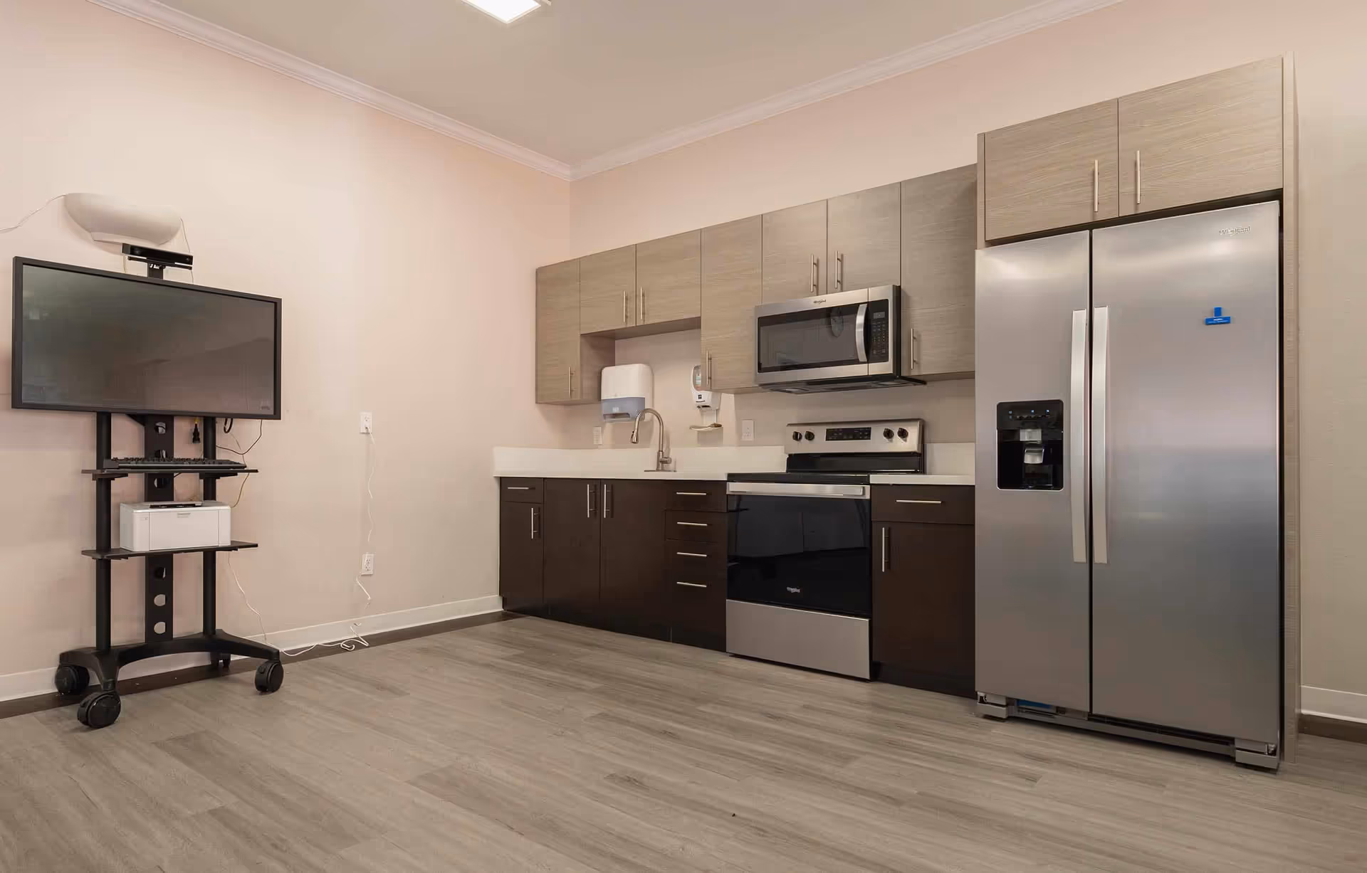 Modern kitchenette with stainless steel refrigerator, stove, microwave, sink, wood cabinets, and a TV on a rolling cart.