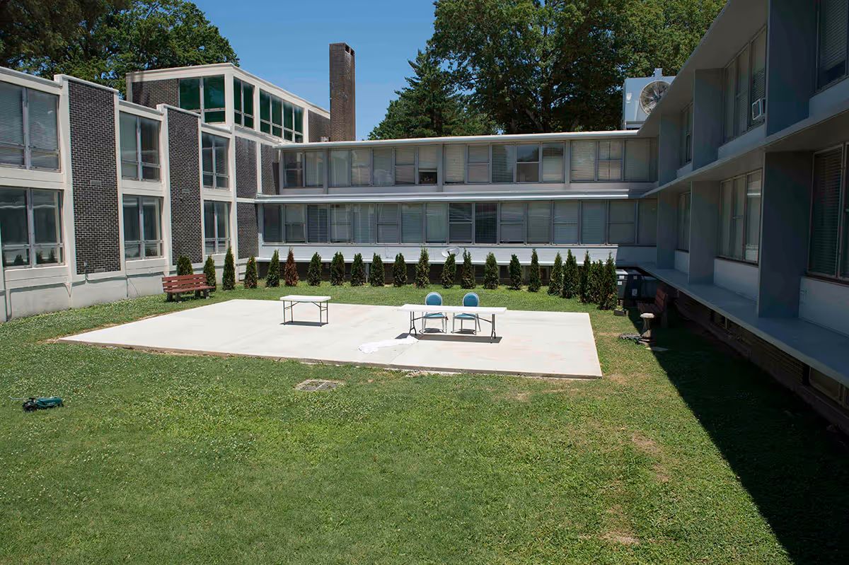 Outdoor courtyard area of a senior living facility with a concrete patio in the center, two blue chairs and a table on the patio, surrounded by green grass and small shrubs. The building has multiple windows and is two stories tall, with a clear blue sky above.