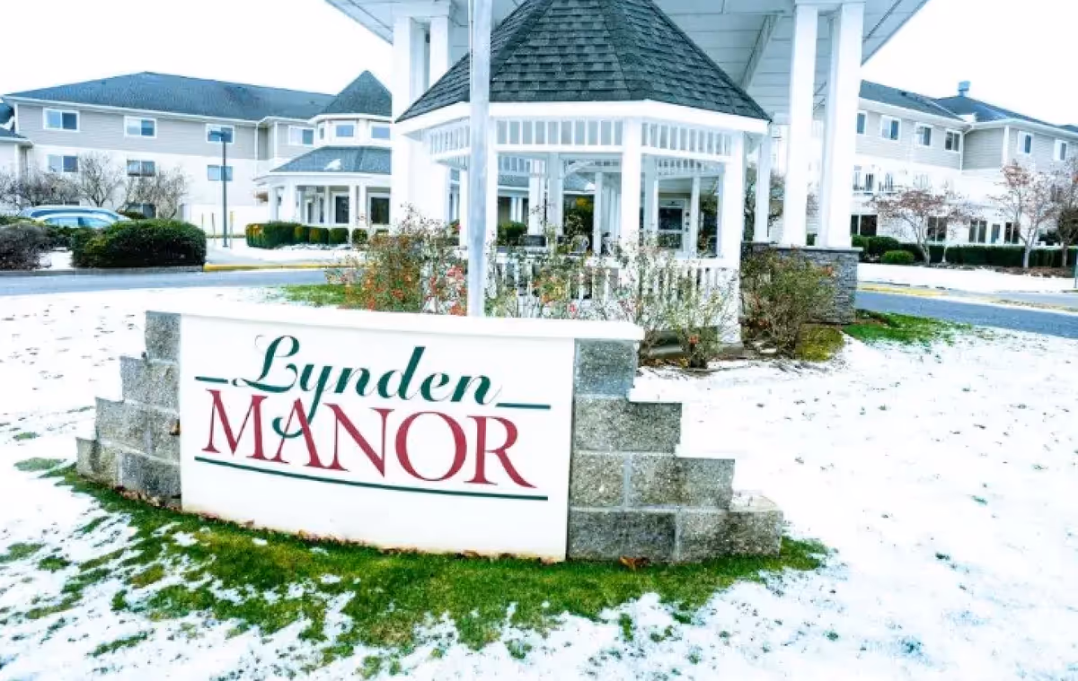 Outdoor view of Lynden Manor senior living facility entrance with a sign displaying the name. The area is lightly covered with snow, and there is a white gazebo and multi-story buildings in the background.