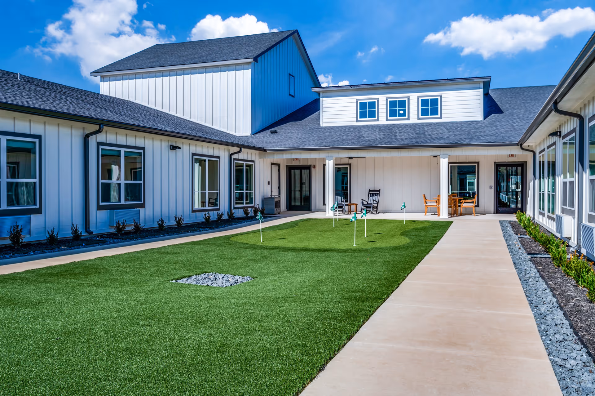 Outdoor courtyard area of The Lodge of Saginaw Health & Wellness featuring a putting green with small flags, surrounded by a concrete walkway and white buildings with multiple windows under a blue sky with scattered clouds.