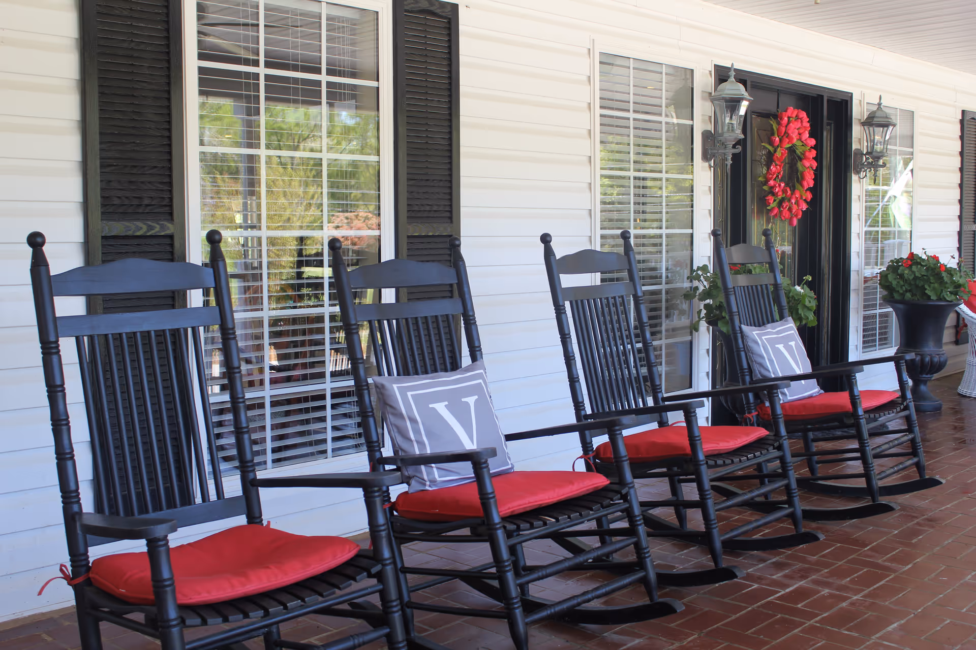 A row of black wooden rocking chairs with red cushions and gray pillows with the letter 'V' on a covered porch with white siding and black shutters. There are large windows with white blinds, a black door decorated with a red wreath, and potted plants on the porch.