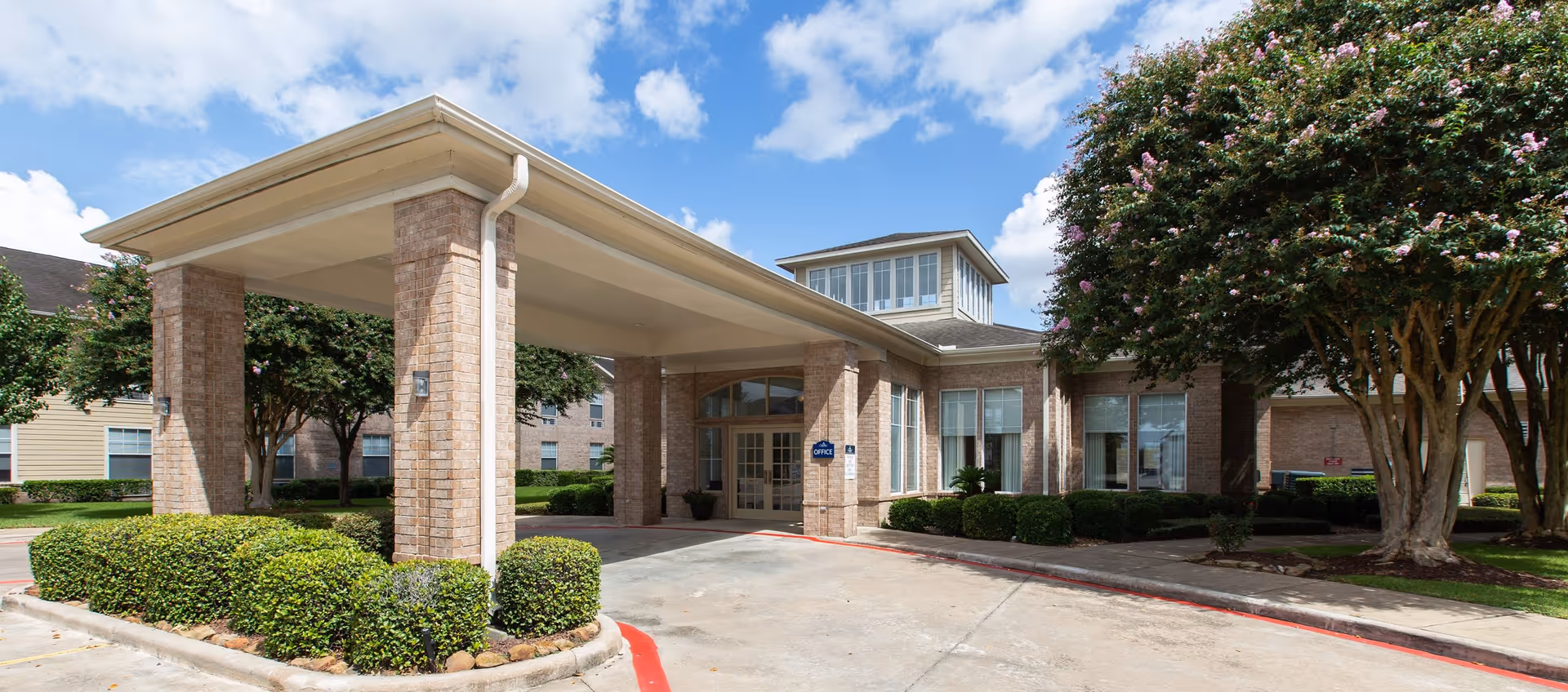 Exterior view of Morada Deer Park facility entrance with a covered drop-off area supported by brick columns, surrounded by neatly trimmed bushes and trees under a partly cloudy blue sky.