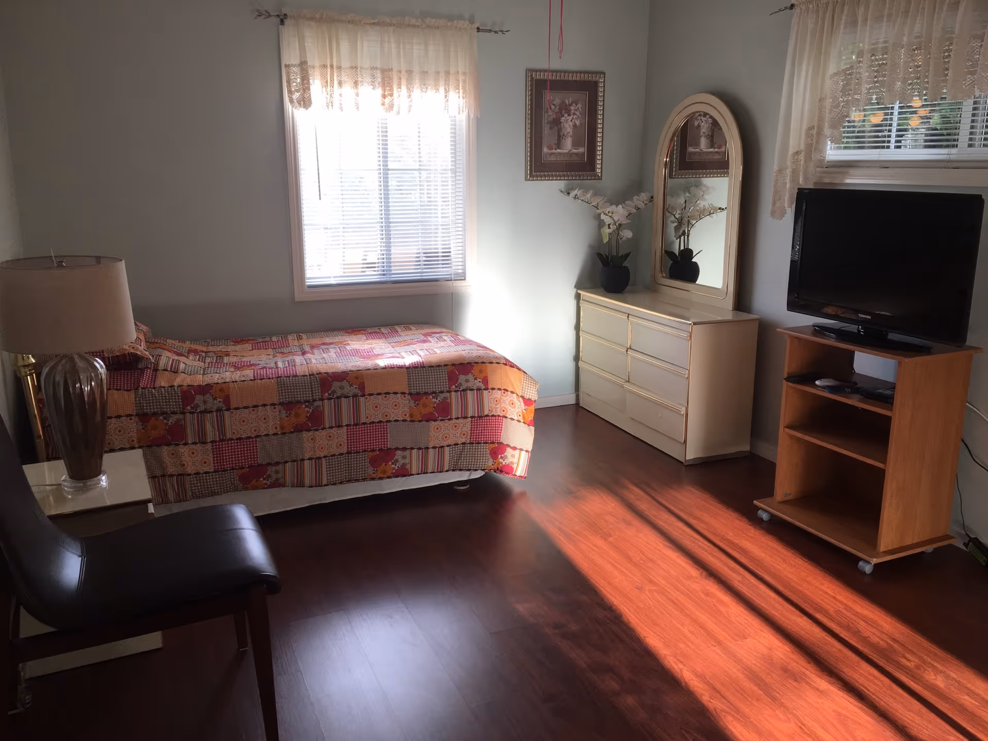 Small bedroom with a single bed, dresser and mirror, TV on a stand, chair and lamp with sunlight on the wood floor.