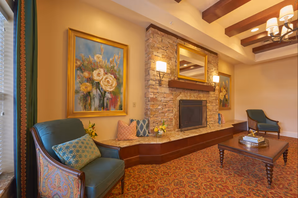 A cozy living room area with two upholstered armchairs, a wooden coffee table with books and decorative items, a stone fireplace with a large mirror above it, and floral paintings on the walls. The room features warm lighting, a patterned carpet, and exposed wooden ceiling beams.