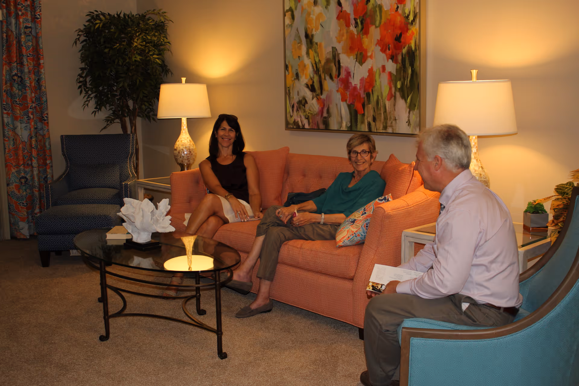 Three people sitting and chatting in a warmly lit living room with a coral sofa, armchairs, lamps, and a glass coffee table.