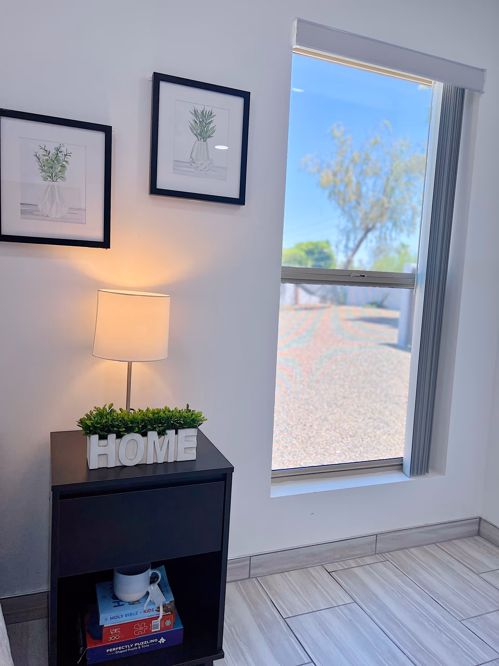 Bright interior corner with a lamp on a black side table topped with a 'HOME' sign and faux greenery, framed botanical prints on the wall, and a tall window looking outside.