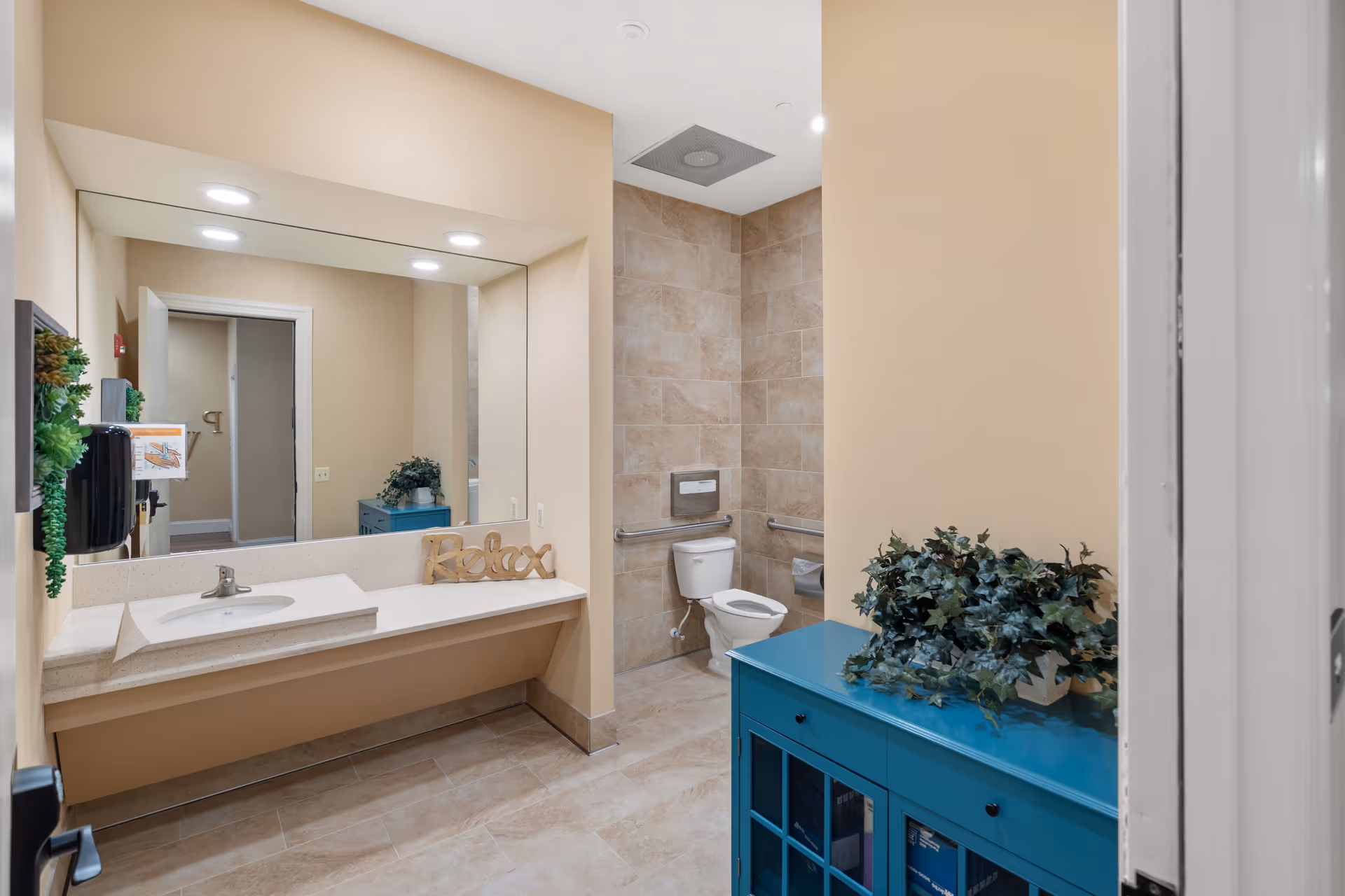 A clean and well-lit bathroom with beige walls and tiled floor. The room features a large mirror above a long countertop with a sink, a decorative 'Relax' sign, and a soap dispenser mounted on the wall. There is a toilet with grab bars on the tiled wall behind it, and a blue cabinet with a green leafy plant on top.
