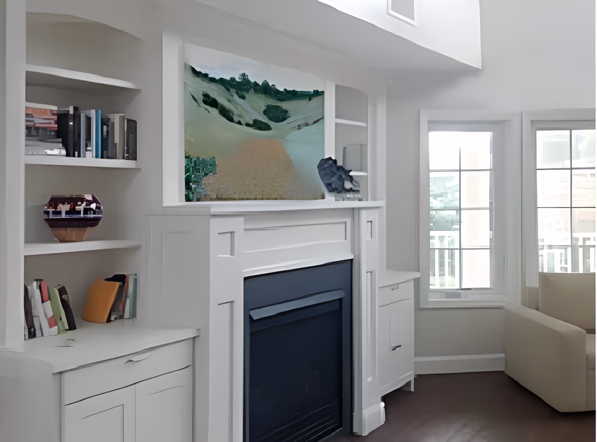 Bright living room interior with a white fireplace mantel and built-in shelving, wall art above the mantel, large windows, and a beige sofa.