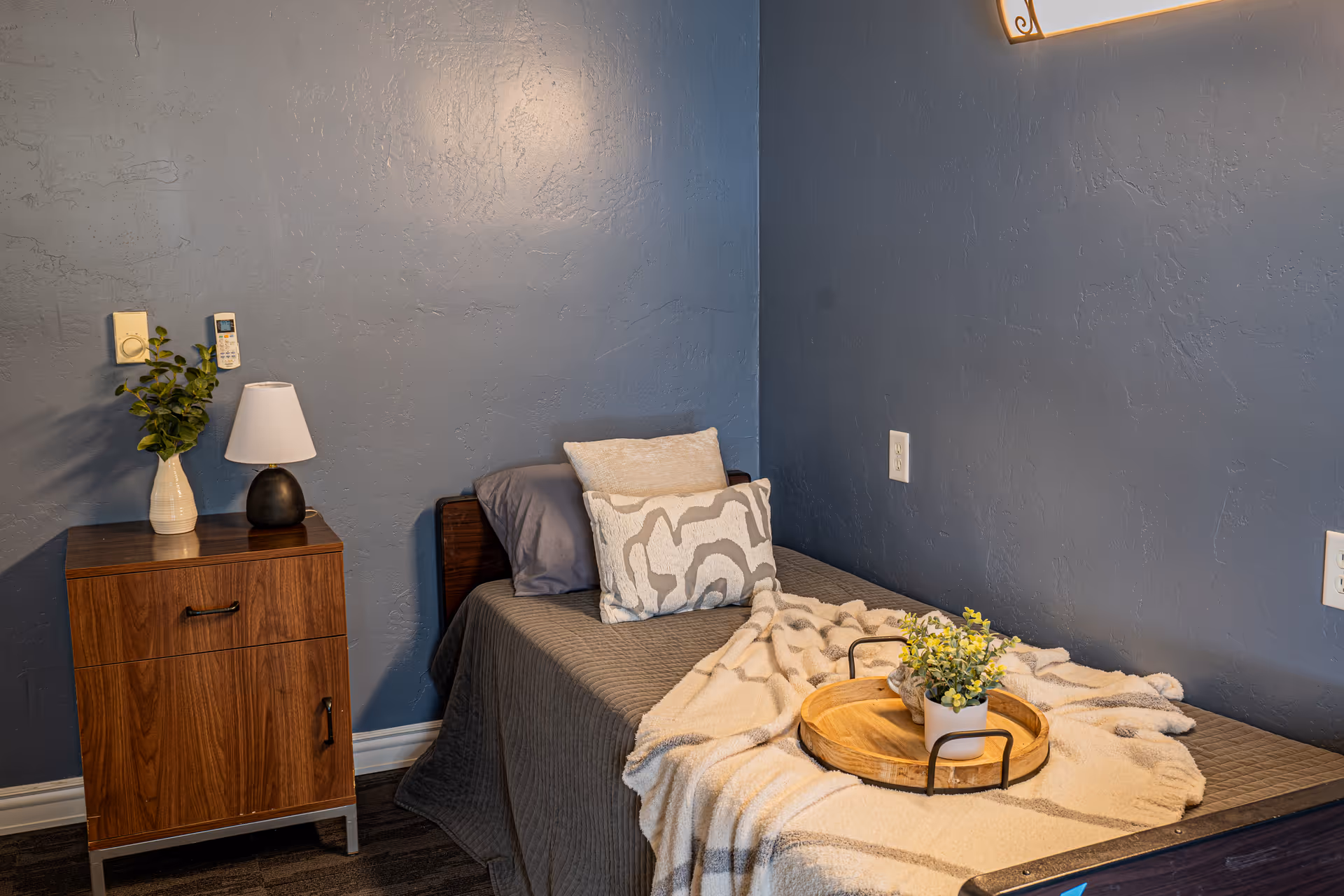 A cozy bedroom with a single bed against a blue textured wall. The bed is made with gray bedding, two decorative pillows, and a beige throw blanket. A wooden nightstand beside the bed holds a small lamp and a vase with green foliage. A wooden tray with a small potted plant is placed on the bed.