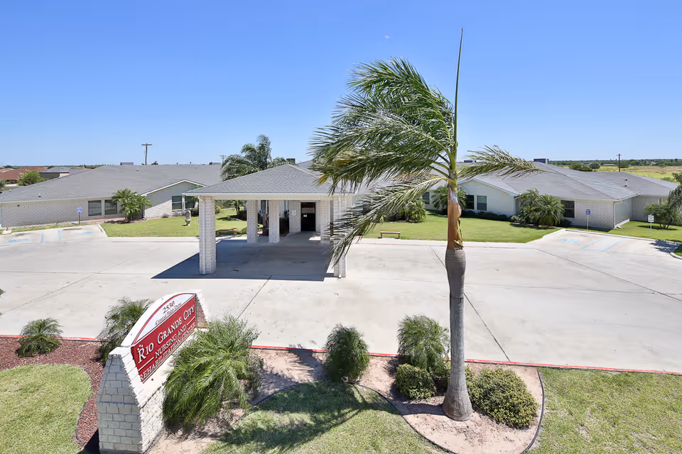 Covered entrance and front driveway of a single-story nursing and rehabilitation center with palm trees and a sign in the landscaped foreground.