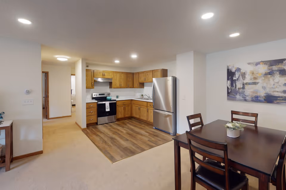 Open-concept kitchen with wooden cabinets, stainless steel appliances and an adjacent dining table in a senior living apartment.