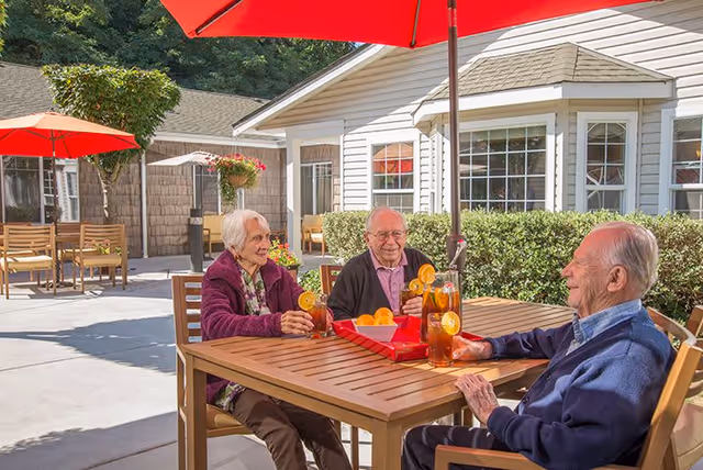 Three elderly people sitting around a wooden outdoor table under a red umbrella, enjoying drinks with lemon slices on a sunny patio outside a residential building with white siding and large windows.