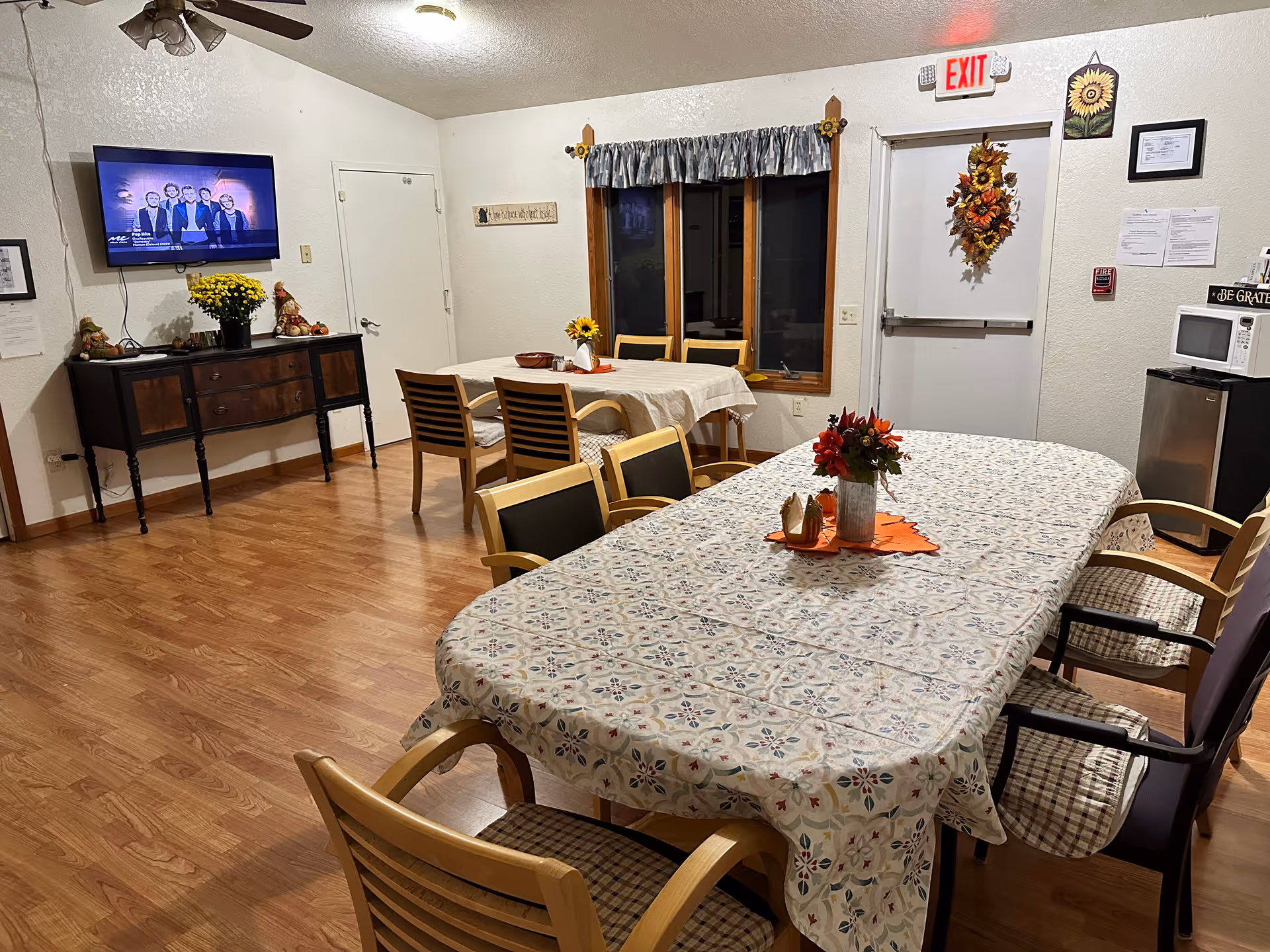 A cozy dining room in an assisted living facility with two tables covered with patterned tablecloths. Each table has chairs around it and a floral centerpiece. There is a wall-mounted TV showing a group of people, a sideboard with decorations, a microwave on a small refrigerator, and an exit door decorated with a fall-themed wreath. The room has wooden flooring and a ceiling fan.