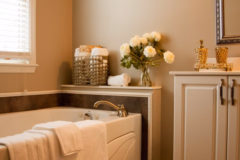 A bathroom corner featuring a white bathtub with a folded white towel draped over the edge. Next to the tub is a shelf holding a basket filled with rolled white towels and a glass vase with white flowers. A cabinet with a decorative gold soap dispenser and matching cup is also visible, along with a window with white blinds letting in natural light.