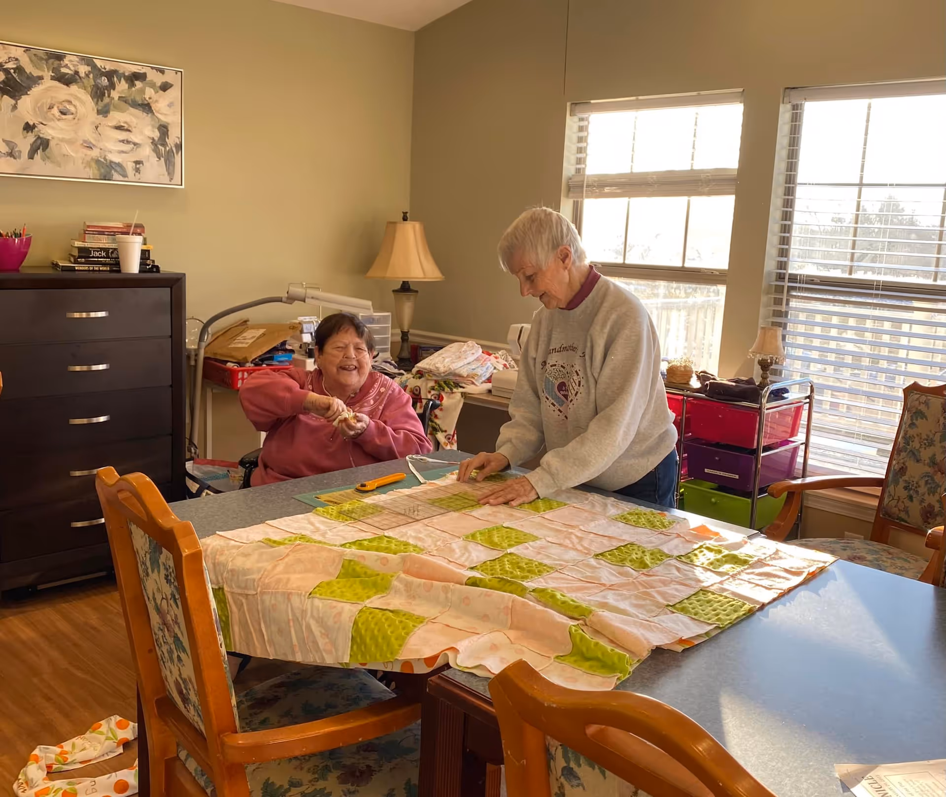 Two elderly women engaged in quilting together at a table in a well-lit room with large windows. One woman is sitting and smiling while holding a quilting tool, and the other is standing and working on the quilt. The room has wooden floors, a dark dresser, a lamp, and floral-patterned chairs.