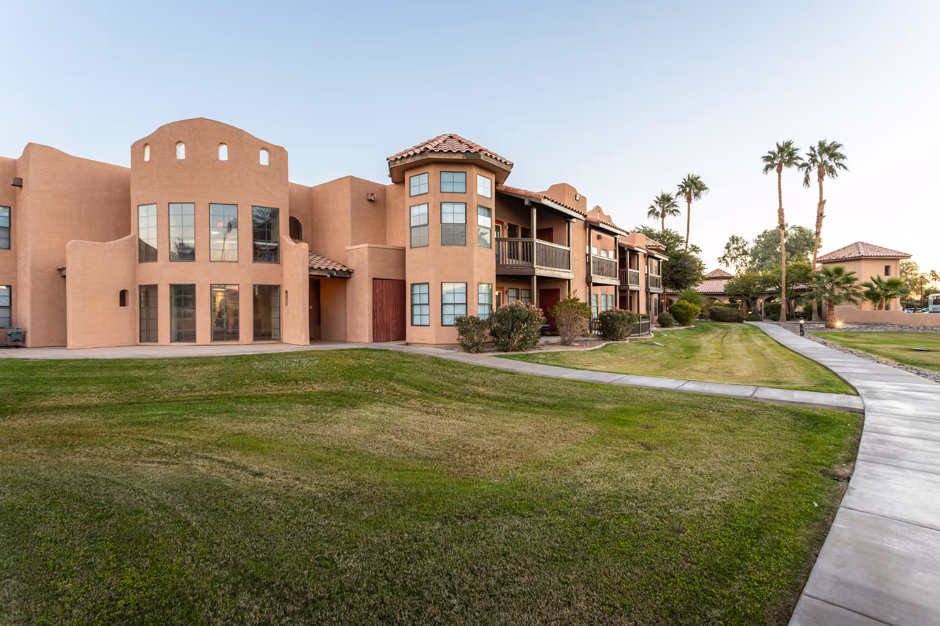 Stucco Mediterranean-style senior living building with balconies, palm trees, and a paved walkway across manicured lawns.