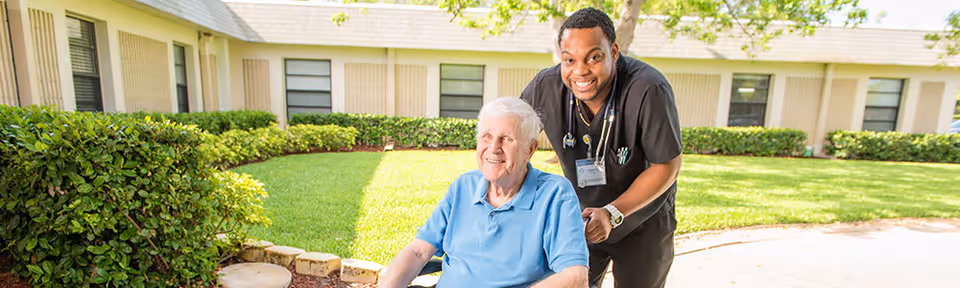 An elderly man in a wheelchair is outdoors on a sunny day, accompanied by a smiling healthcare worker standing behind him. They are in front of a building with windows and surrounded by green bushes and a well-maintained lawn.
