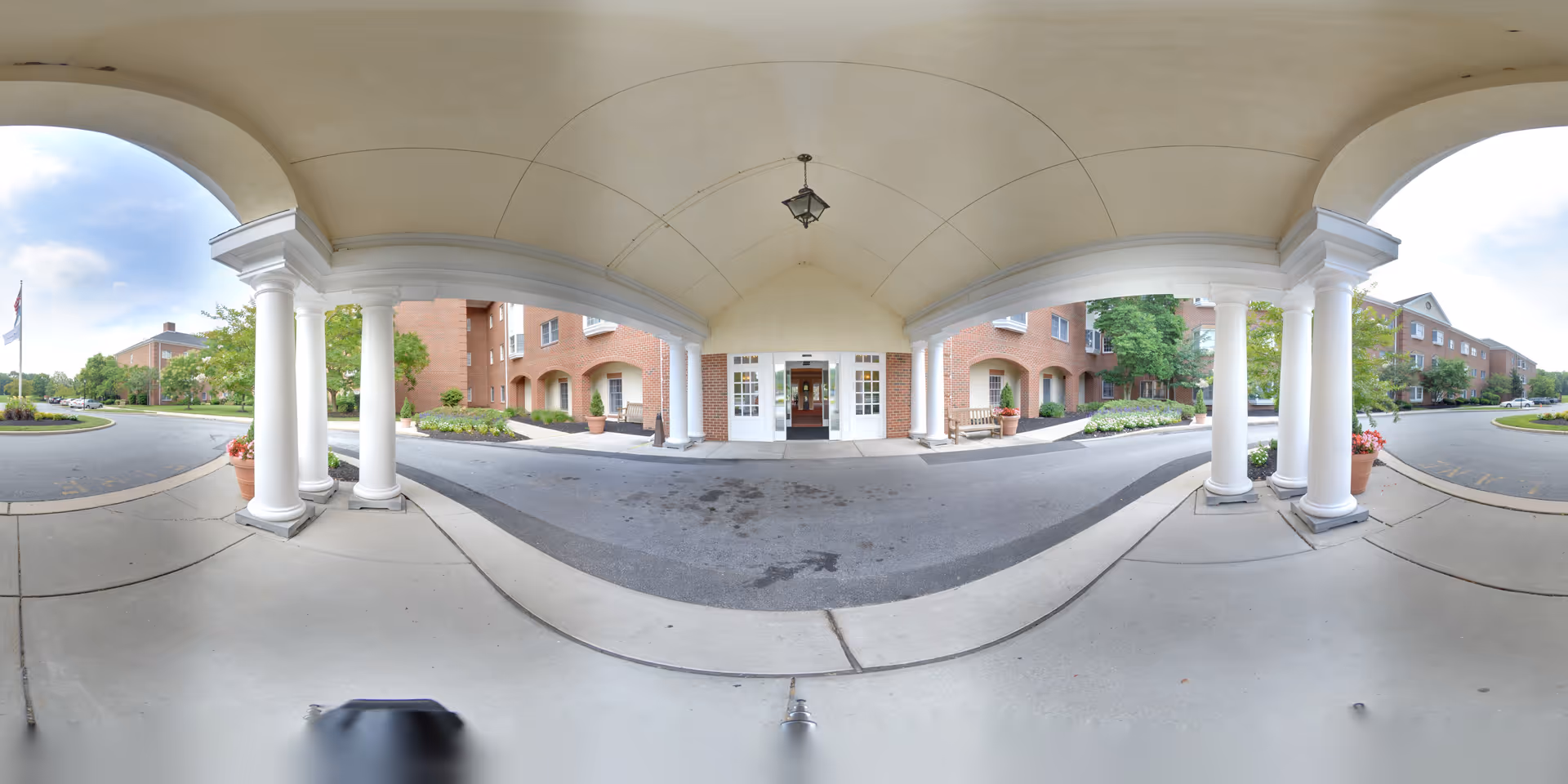 Covered entrance area of a senior living facility with white columns supporting a curved roof. The entrance has double glass doors leading into a brick building. There are landscaped areas with plants and trees on either side of the driveway, and the sky is partly cloudy.