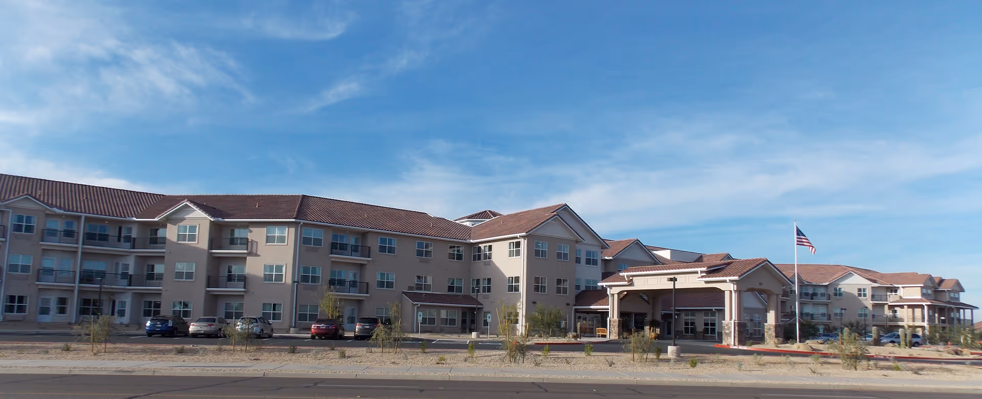 Wide exterior view of a large, multi-story senior living facility with beige walls and red-tiled roofs under a blue sky with some clouds. The building has multiple balconies and an American flag flying near the entrance. Several cars are parked in front of the building.