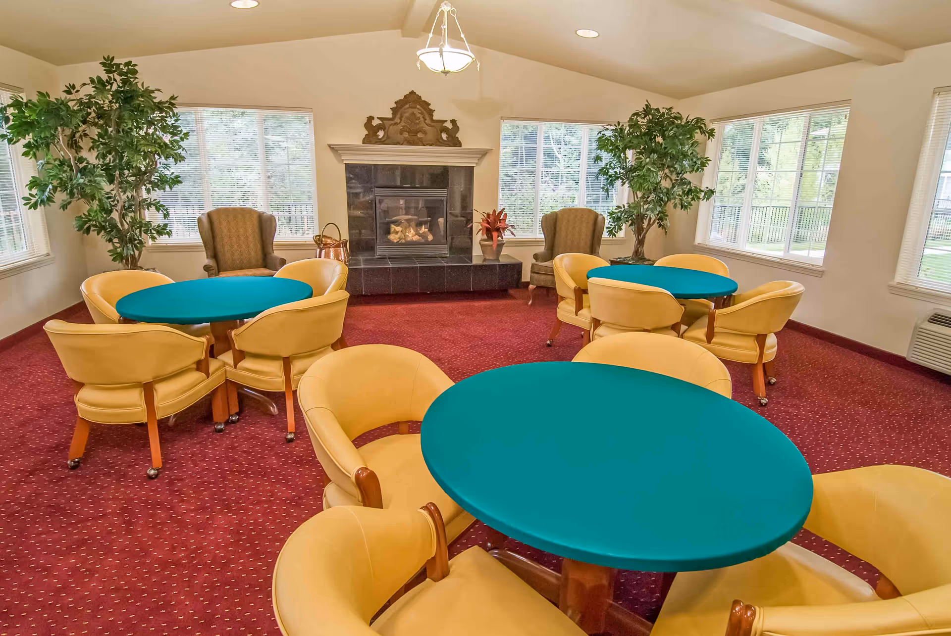 A cozy common room with three round tables covered in teal tablecloths, each surrounded by yellow cushioned chairs. The room has large windows letting in natural light, two large potted plants, two armchairs near a black tiled fireplace with a decorative wooden mantle, and a red carpeted floor.