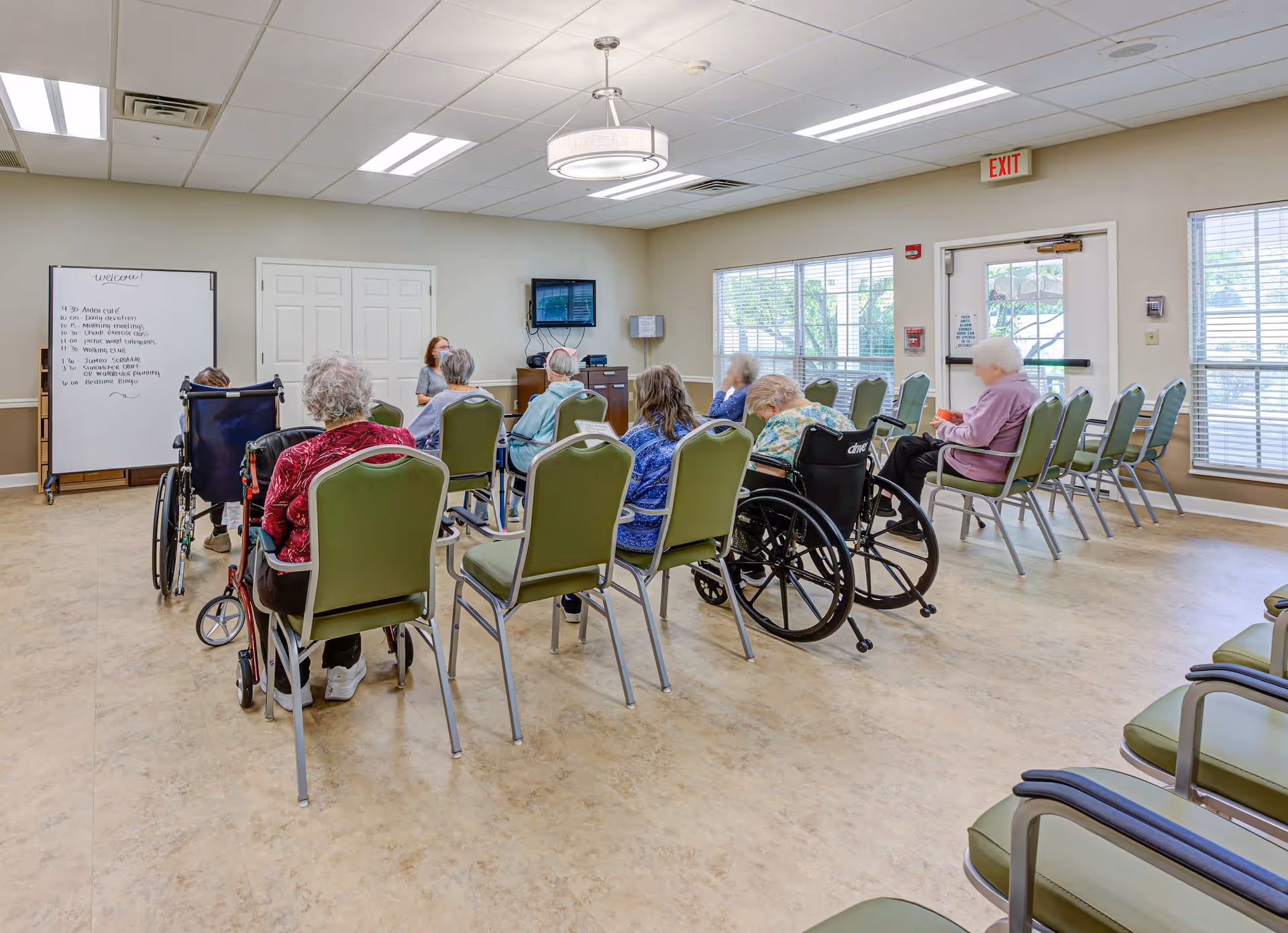 A group of elderly individuals, some in wheelchairs, seated in a semi-circle in a bright, spacious room with beige walls and large windows. A caregiver or staff member is seated facing them near a whiteboard with a schedule written on it. The room has green chairs, a wall-mounted TV, and an exit door with windows letting in natural light.
