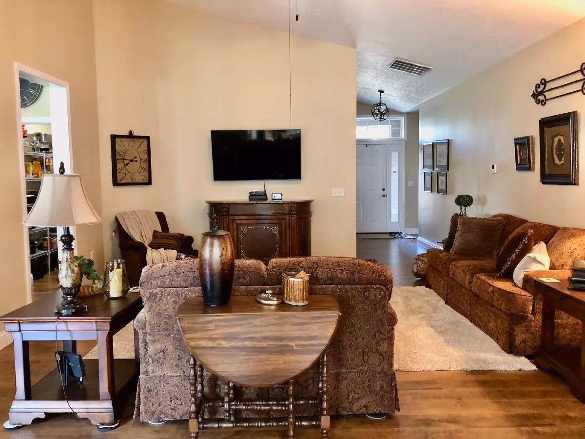 A cozy living room with two patterned brown sofas, a wooden side table with a lamp and decorative items, a small wooden drop-leaf table with vases, a wall-mounted flat-screen TV above a wooden cabinet, and a hallway leading to a white front door with sidelights. The walls are beige, and there are framed pictures and a clock on the walls.