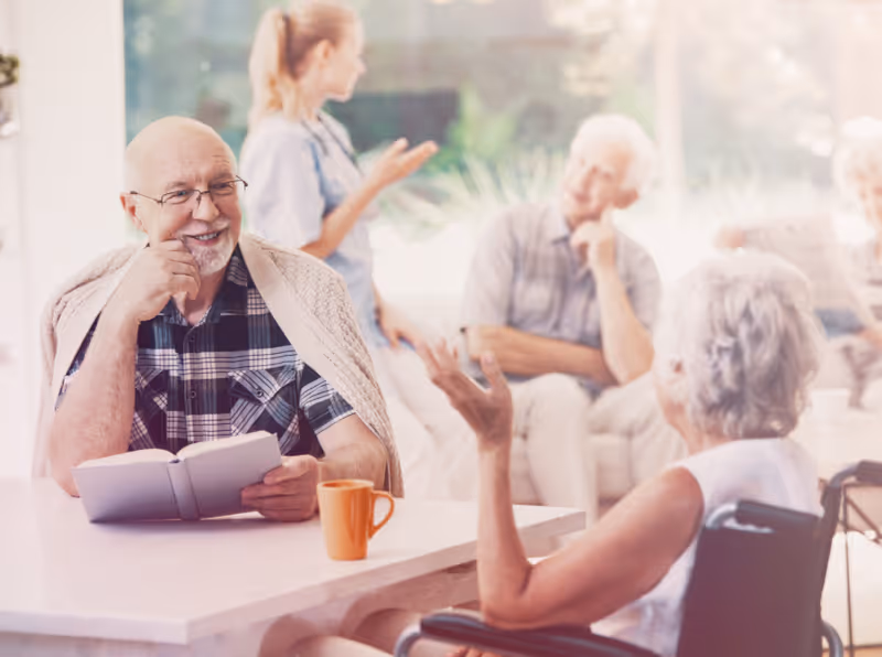 A smiling elderly man reads a book at a table while other residents and a caregiver chat in a bright communal lounge.