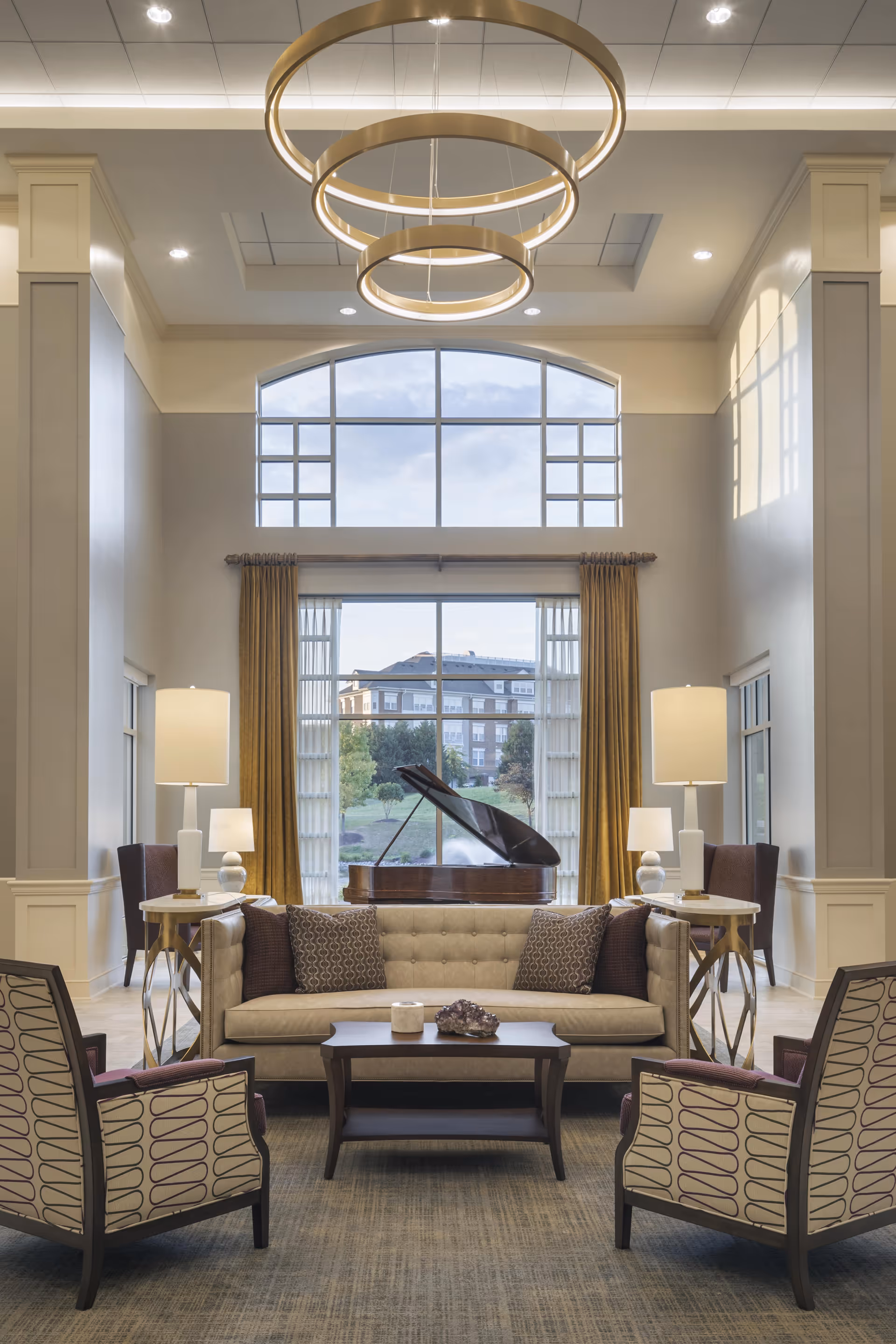 A spacious and elegant living room with high ceilings, large windows with golden curtains, and a grand piano near the window. The room features a beige tufted sofa with patterned cushions, two patterned armchairs, a wooden coffee table with decorative items, and two side tables with lamps. Modern circular chandeliers hang from the ceiling, and natural light fills the room.