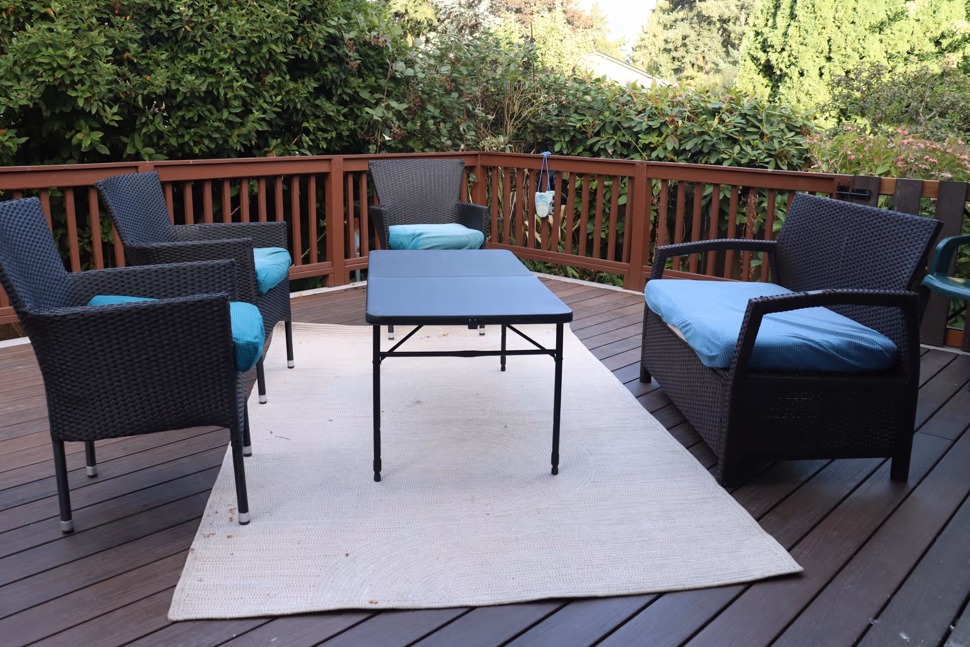 Outdoor wooden deck with a rectangular black table surrounded by four black wicker chairs and one black wicker loveseat, all with light blue cushions. The deck is enclosed by a wooden railing and surrounded by green bushes and trees.