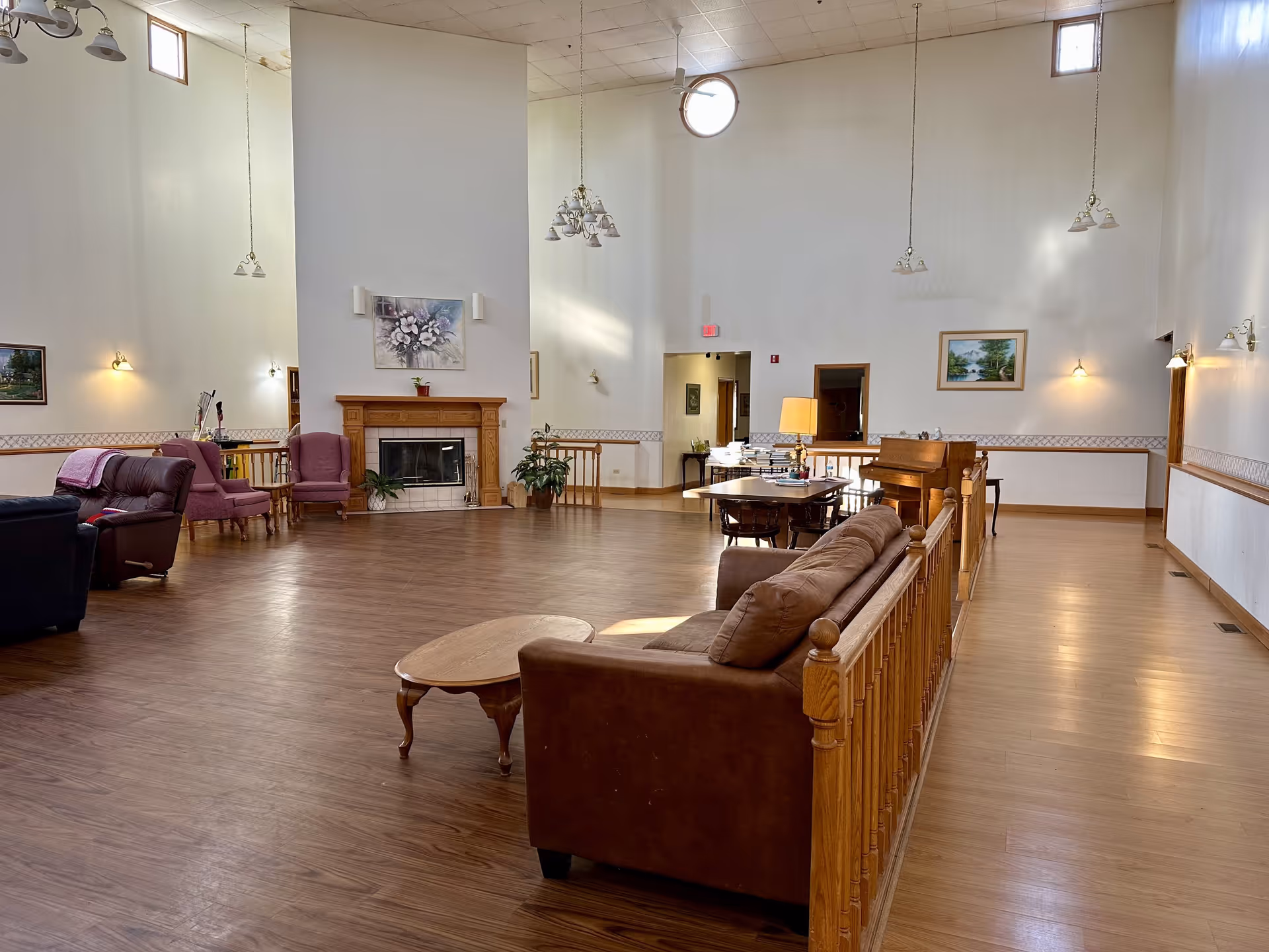 A spacious living room area with high ceilings and wooden flooring. The room features a brown sofa, a wooden coffee table, a fireplace with a floral painting above it, several armchairs, a piano, and a dining table with chairs. The walls are light-colored with small windows near the ceiling allowing natural light to enter. Multiple hanging light fixtures and wall sconces provide additional lighting.