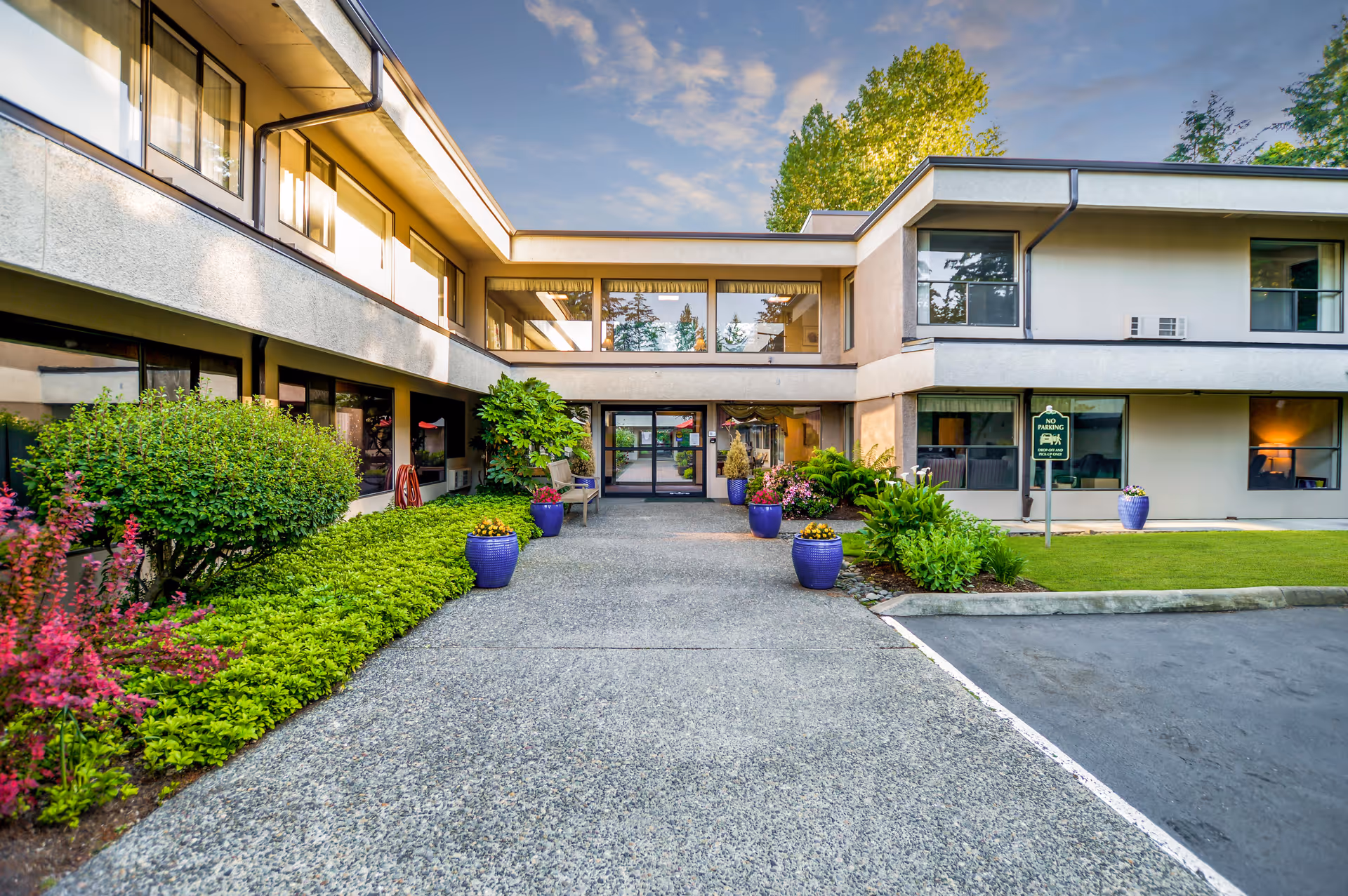 Exterior view of a two-story assisted living facility building with large windows, a paved walkway lined with green shrubs and potted plants, and a clear sky with some clouds.