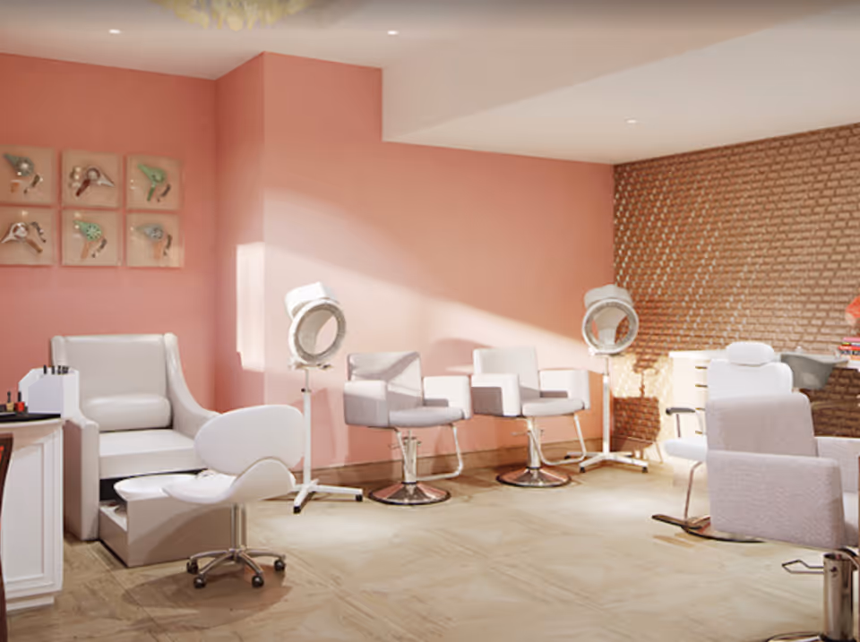 A bright and modern salon area with pink walls and a textured brown accent wall. The room features several salon chairs, including white and light gray styling chairs, two white hair dryers on stands, and a white pedicure chair with a matching footrest and stool. There is also a small table with nail polish bottles and a decorative wall art piece with bird designs.