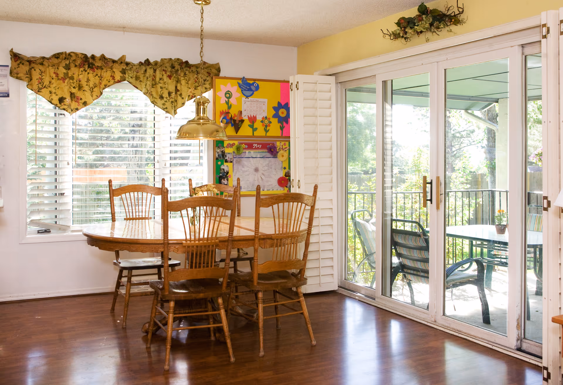 A dining area with a wooden table and six wooden chairs on a hardwood floor. A brass pendant light hangs above the table. There is a large window with white blinds and a floral valance on the left wall. On the right side, there are sliding glass doors leading to an outdoor patio with chairs and a table. A colorful bulletin board with artwork and notes is mounted on the wall near the window.