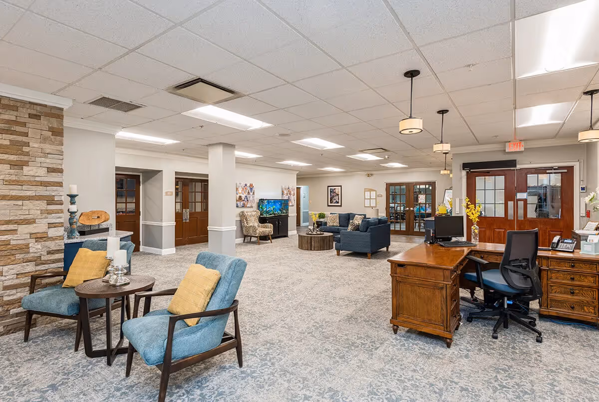 A spacious and well-lit common area in a senior living facility featuring a wooden reception desk with a computer and office chair, two blue armchairs with yellow cushions around a small round table, a seating area with a blue sofa and patterned armchair, a fish tank, and multiple wooden doors with glass panels. The room has a light gray carpet and a drop ceiling with fluorescent lights and pendant lamps.