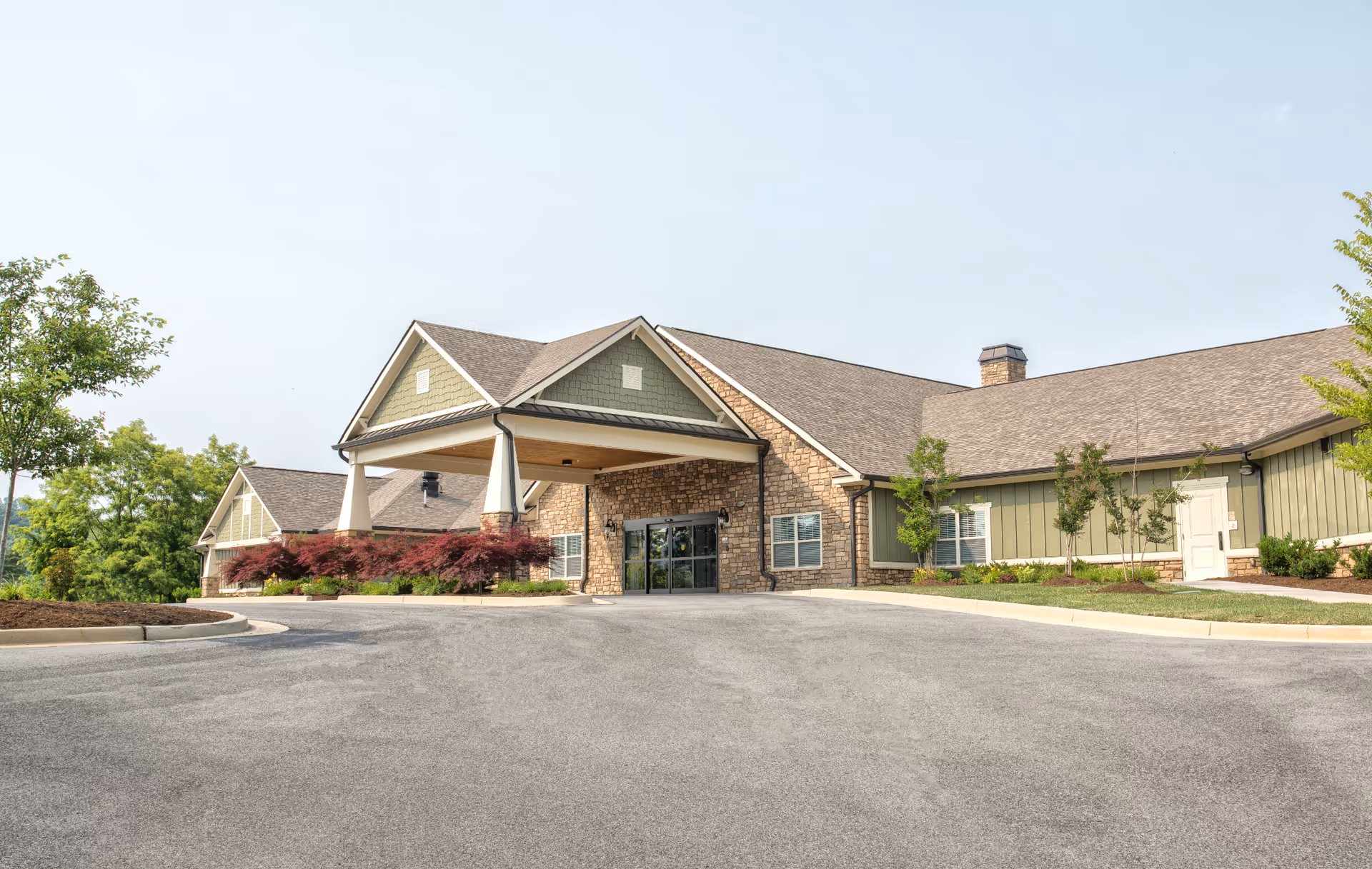 Exterior view of Dominion Senior Living of Bristol building with a covered entrance, stone and green siding, surrounded by landscaped greenery and trees under a clear sky.