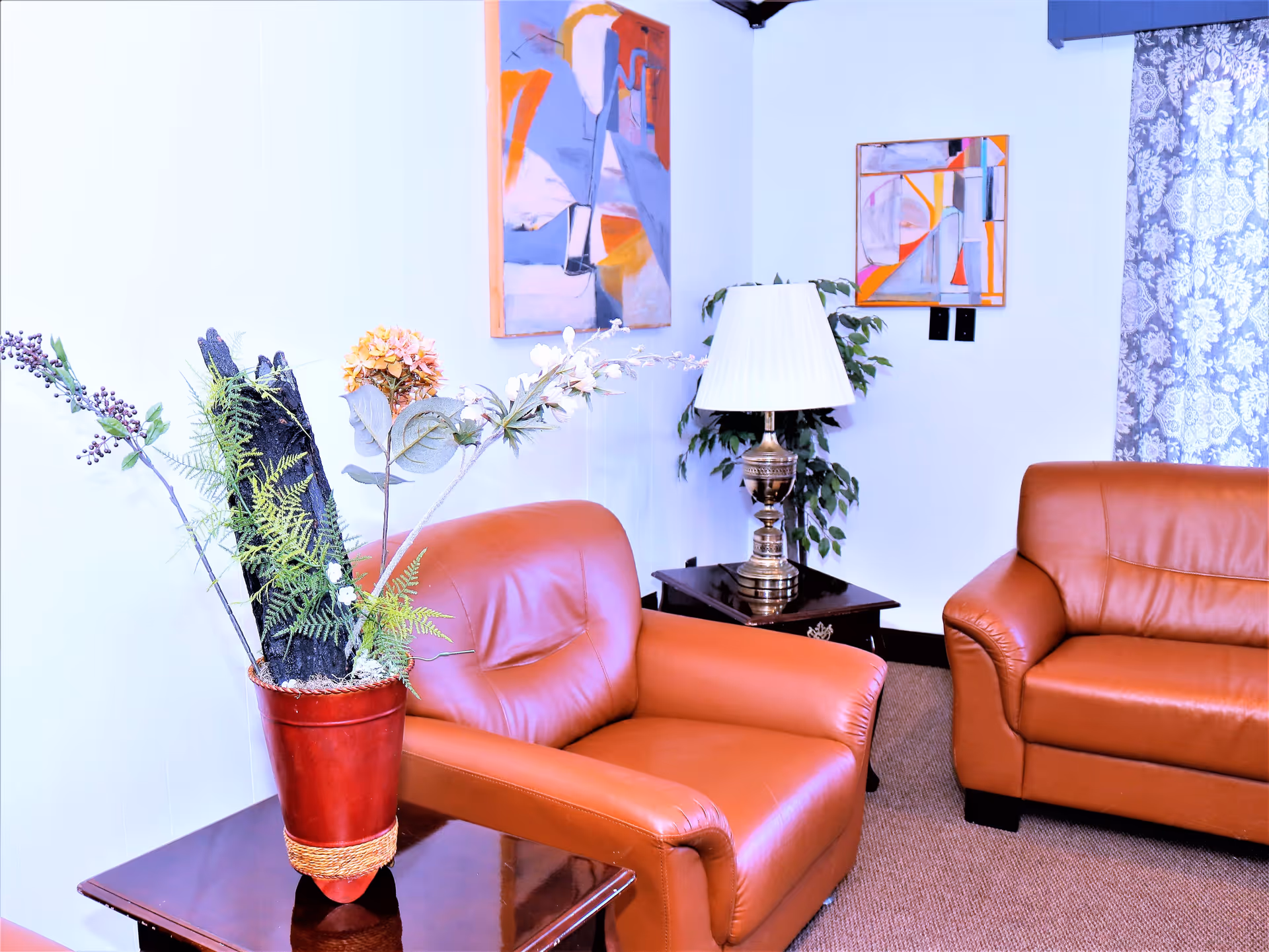 A cozy living room area with two brown leather armchairs and a matching sofa. There is a wooden side table with a decorative flower arrangement in a red vase. Another side table holds a brass lamp with a white lampshade. The walls are white and decorated with two abstract paintings. A window with patterned curtains is partially visible on the right side.