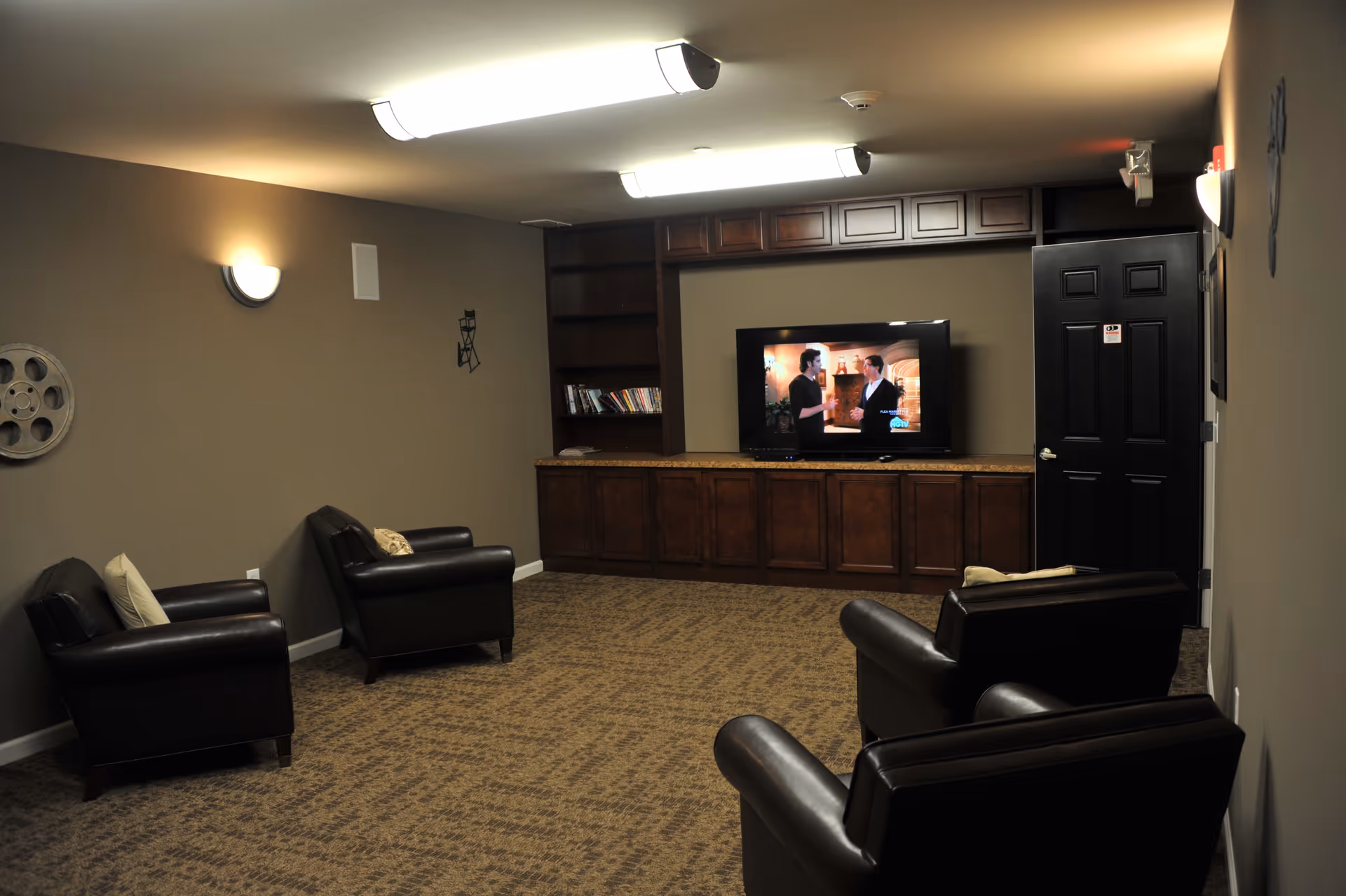 Communal lounge with leather armchairs facing a large television set within built-in wooden cabinets.