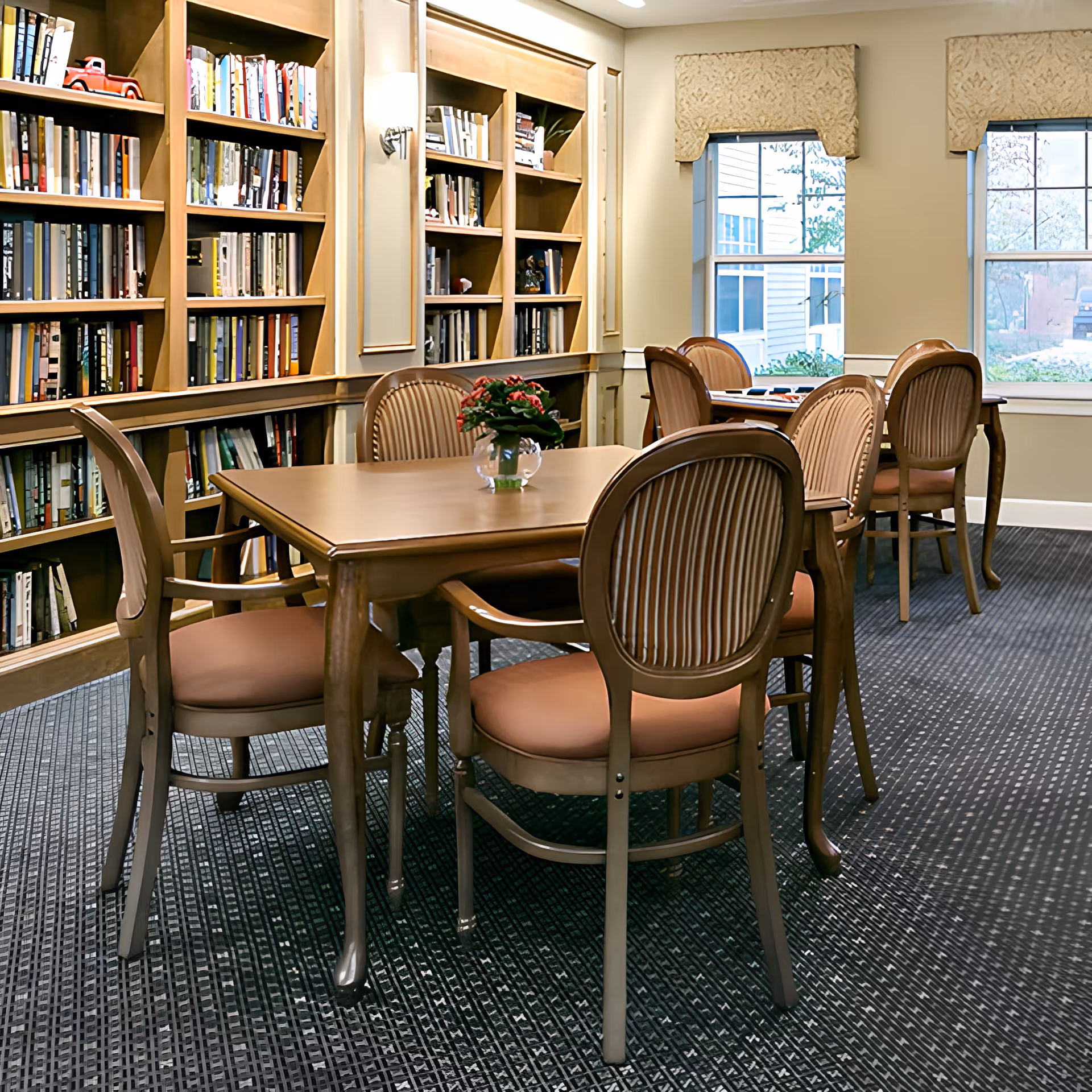 A bright common room with wooden tables and chairs, bookshelves along one wall, and windows letting in daylight.
