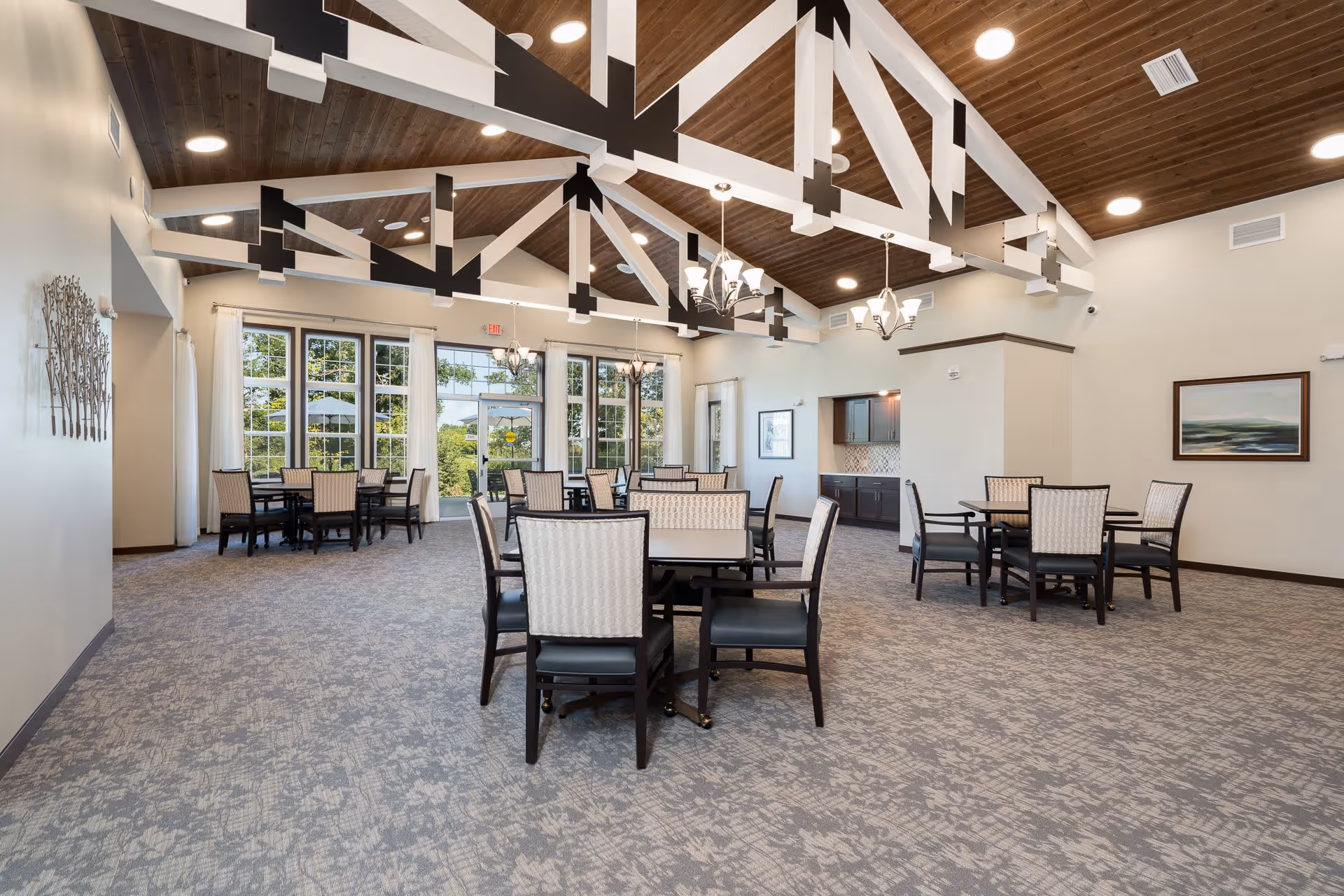 A spacious dining room with multiple tables and chairs arranged neatly. The room features a high wooden ceiling with exposed white and black beams, large windows letting in natural light, and elegant chandeliers hanging from the ceiling. The walls are light-colored with minimal decor, and there is a small kitchenette area with dark cabinets and a tiled backsplash.